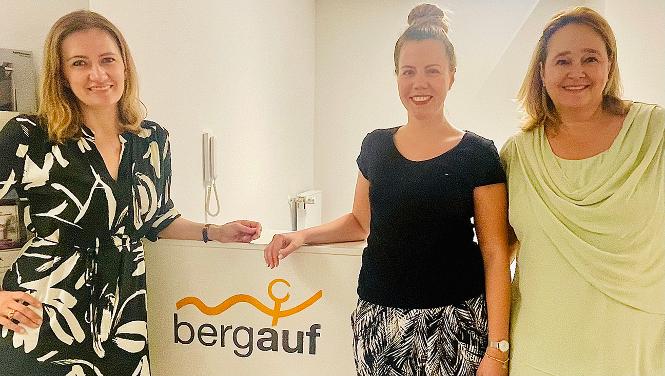 Three women standing together in an office space with a sign that reads 'bergauf'.