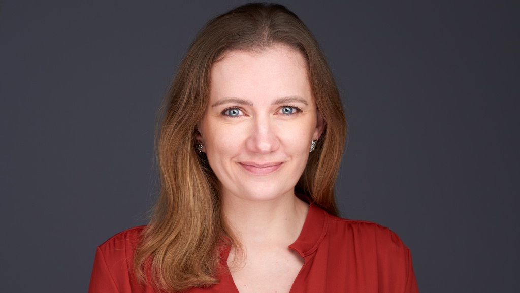A woman with brown hair wearing a red blouse and smiling at the camera against a gray background.