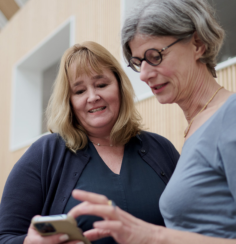 Two women looking at a smartphone together in an indoor setting, one woman smiling and the other focused on the screen.