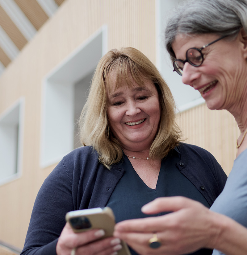 Two women smiling while looking at smartphones in a modern indoor setting.