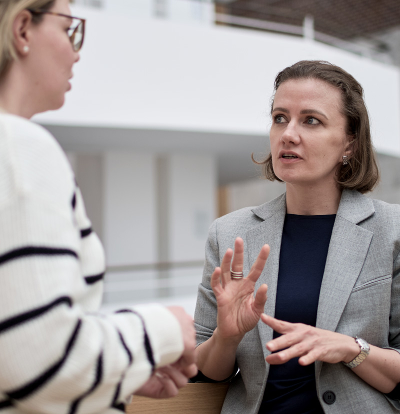 Two women in business attire engaged in a conversation indoors, one gesturing while speaking.
