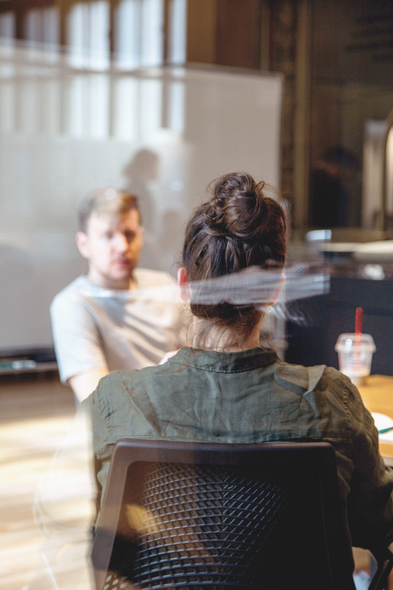 Dieses Foto zeigt eine Frau von hinten, sitzend auf einem Stuhl. Der Fokus des Bildes liegt auf der Frau. Vor der Frau sitzt ein Mann, den man nur unscharf erkennen kann. Die beiden Personen gucken sich an.