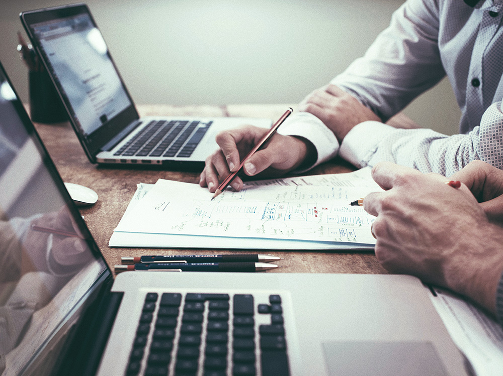 Das Foto zeigt einen Tisch mit zwei Macbook Laptops. Zwischen den zwei Laptops liegt ein Stapel Papier, auf dem viel draufgeschrieben wurde. Vor den Laptops sitzen jeweils zwei Männer, die einen Stift in der Hand haben und beide zeigen mit dem Stift auf den Stapel Papier vor ihnen.