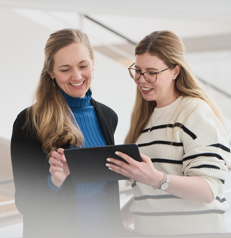 Two women smiling while looking at a tablet in a well-lit indoor space.
