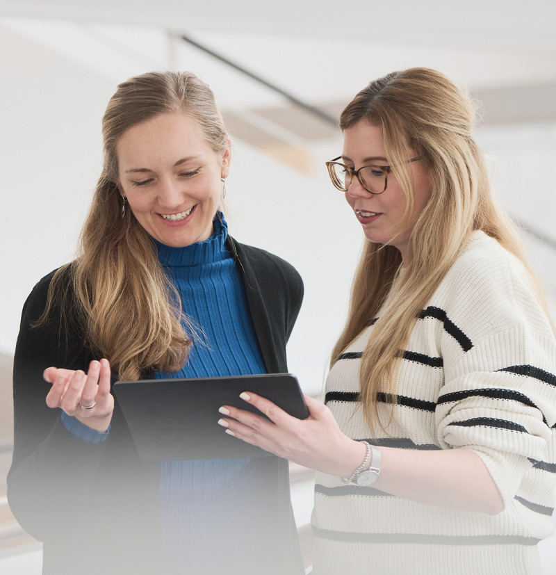 Two women looking at a tablet together, one wearing a blue turtleneck and the other in a striped sweater.