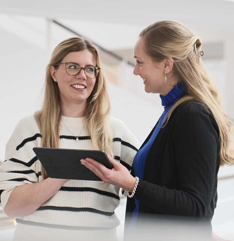 Two women smiling and discussing while holding a tablet in a modern indoor setting.