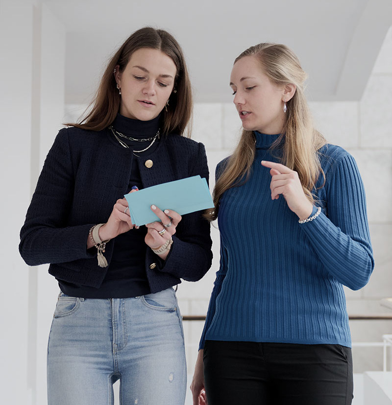 Two women discussing information on a tablet while standing indoors in a modern setting.