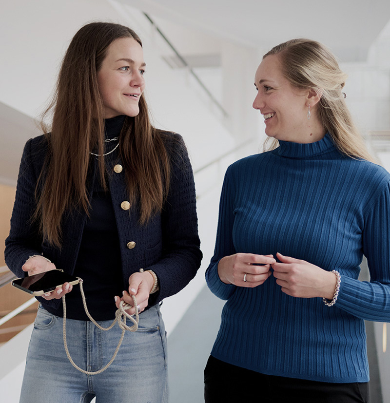 Two women talking while holding a smartphone and smiling at each other in a modern indoor setting.