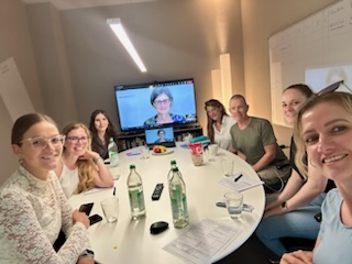 Foto zeigt PULSAR Medical AG Teammitglieder, die an einem ovalen Tisch sitzen und in die Kamera lächeln. Am Ende des Tischs steht ein Fernseher, auf dem Frau Dr. Barbara Heiden zu sehen ist (Videokonferenz).